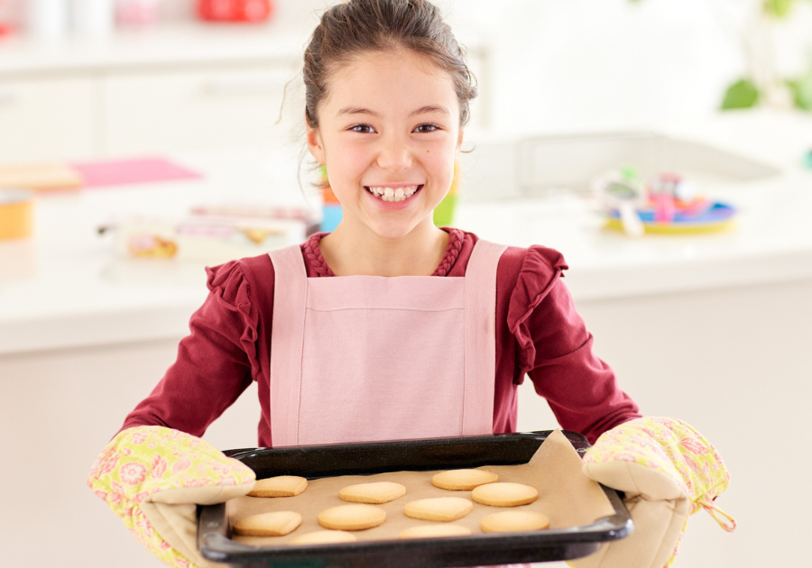 image of a girl standing in the kitchen with oven mittens on holding a baking sheet with cookies on it.