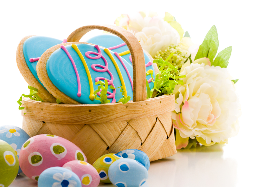 image of an easter basket sitting on a white table filled with colorful decorated easter themed sugar cookies. There are colorful decorated easter eggs and flowers surrounding the easter basket.