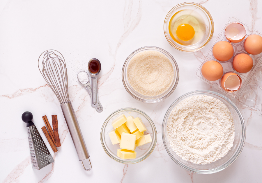 image of flour, sugar, eggs, butter, and baking utensils laying on a marbled white background.